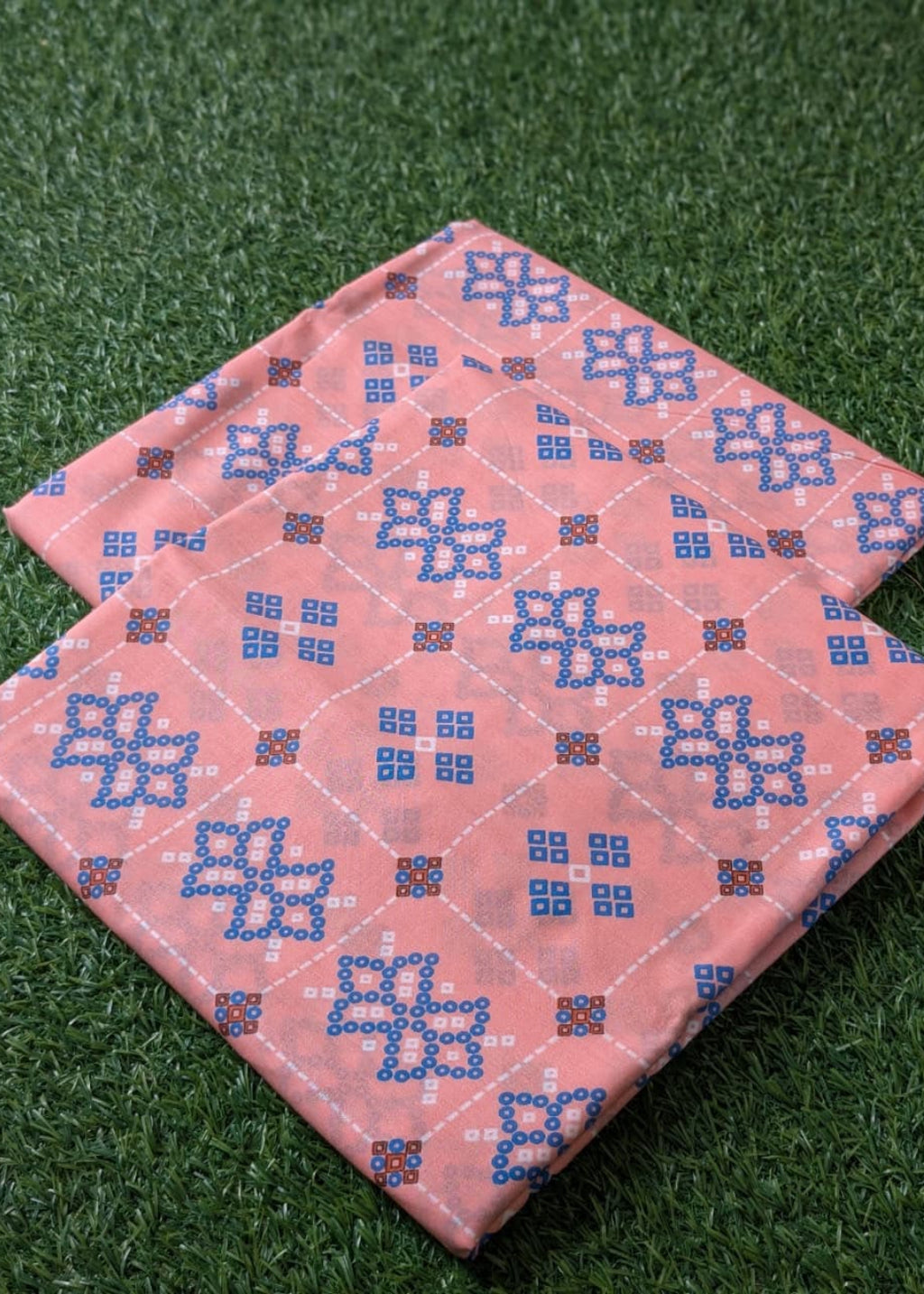 A folded pink fabric item, likely a quilt or blanket, featuring a blue and white geometric pattern, displayed on green artificial grass.