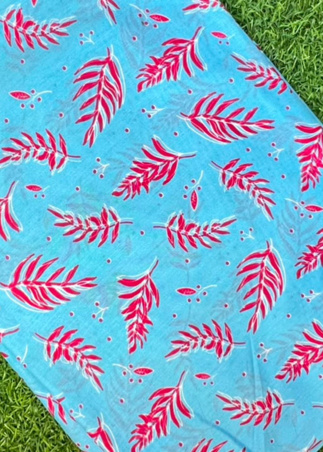 A square fabric item, likely a napkin, small tablecloth, or scarf, featuring a bright blue background with a repeating pattern of pink palm leaves and small white dots, laid flat on green grass.