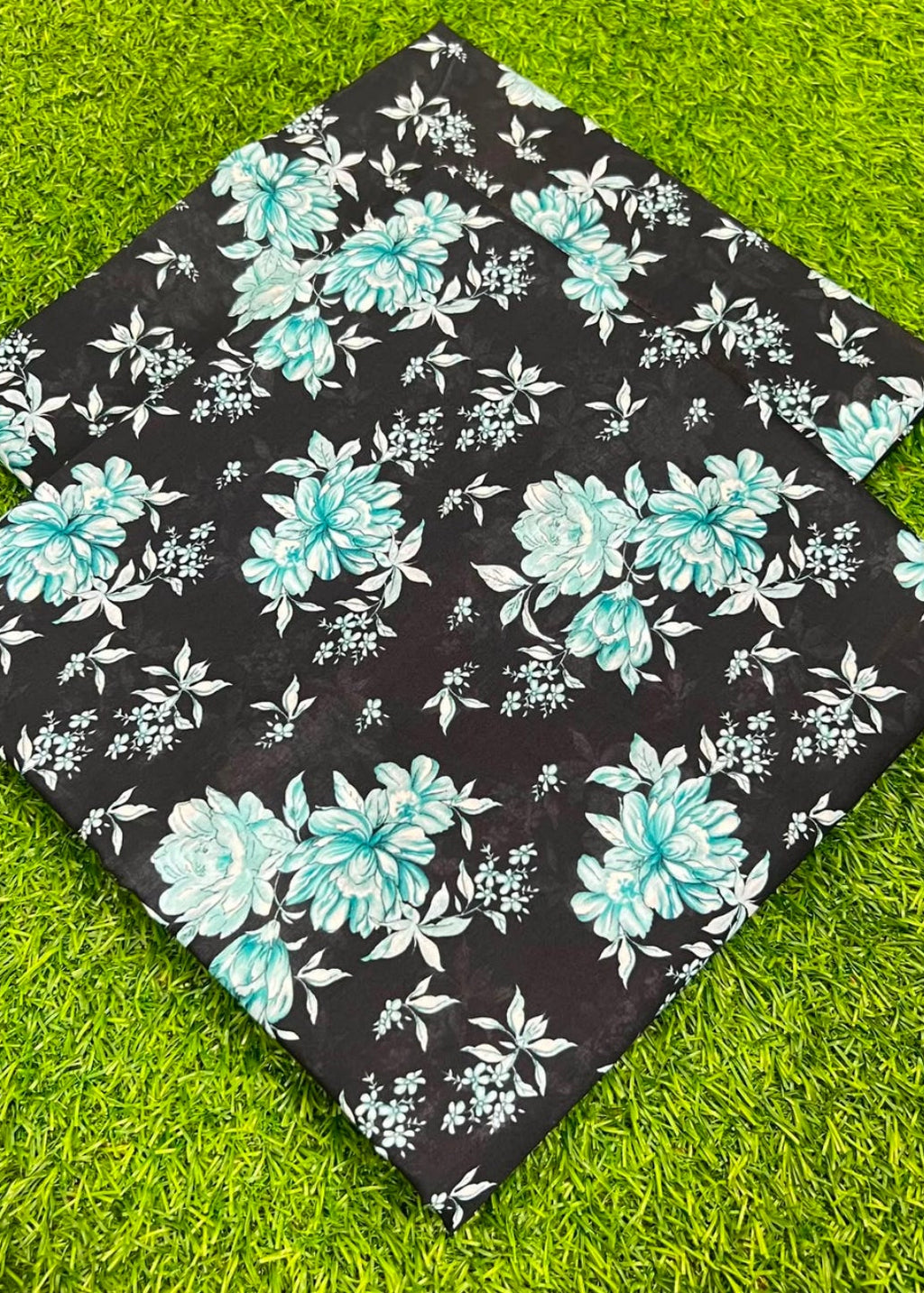 A square black fabric item, likely a quilt or cushion cover, featuring a floral pattern with large teal and light blue flowers and white leaves, laid flat on bright green grass.