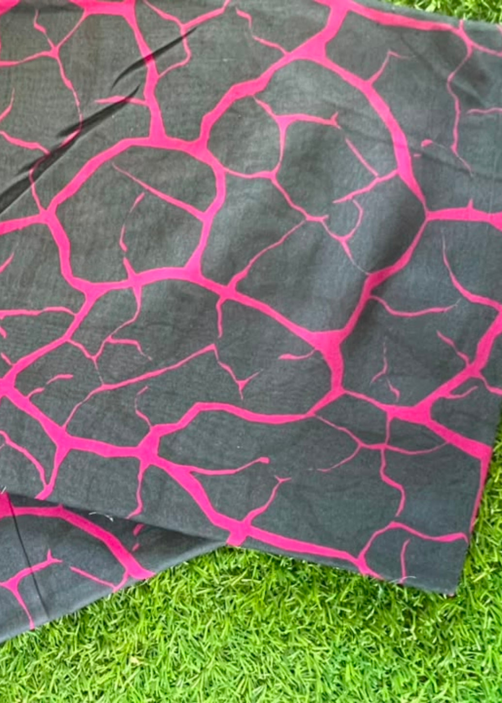 Two square fabric items with a black background and bright pink crackle pattern, laid flat on green grass.
