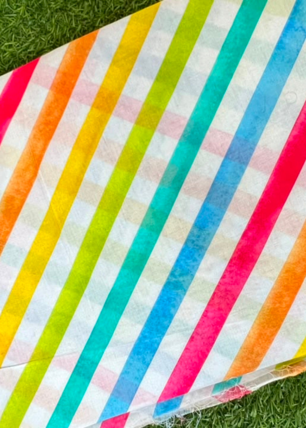 A square fabric item, likely a napkin, coaster, or small mat, featuring a vibrant diagonal striped pattern in pink, orange, yellow, green, and blue on a white background, laid flat on green grass.