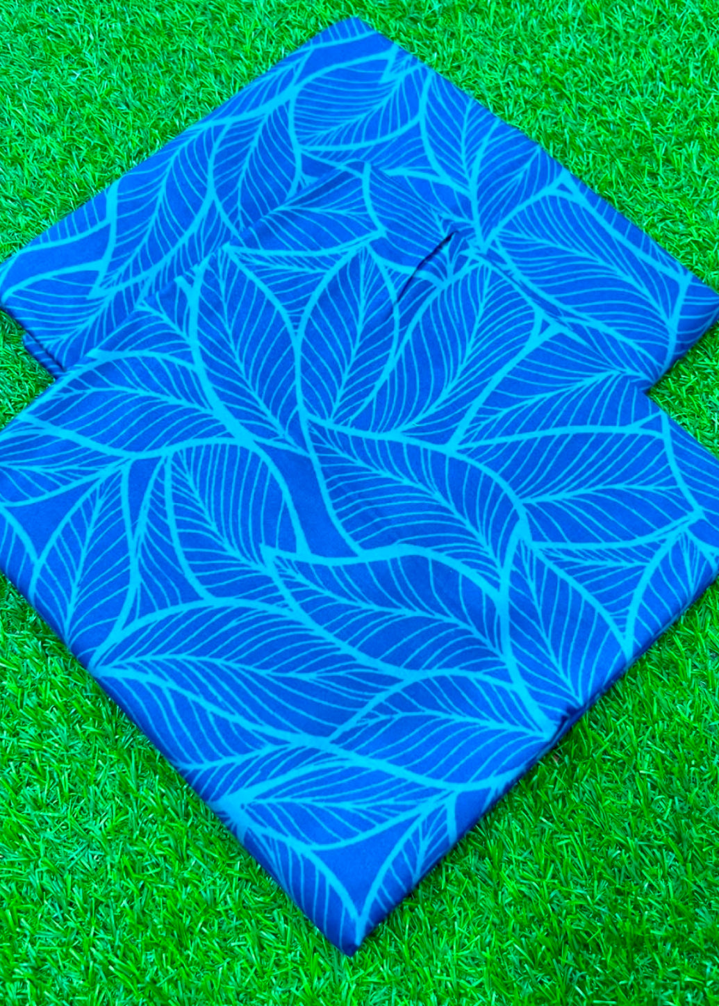 Two square blue outdoor floor cushions with a light blue leaf pattern, displayed on bright green artificial grass.