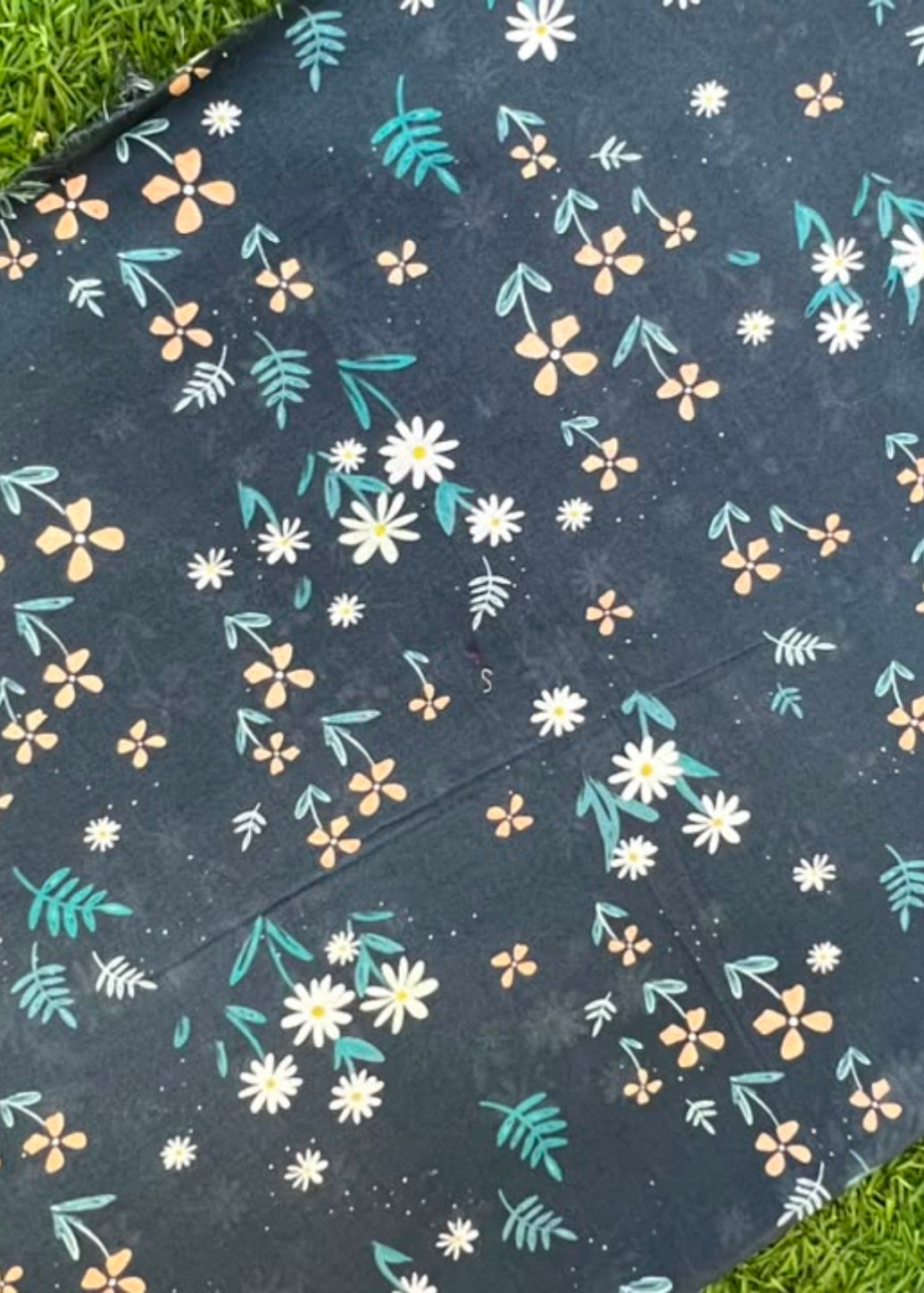 A rectangular fabric item, likely a blanket or mat, featuring a dark navy blue background with a white, yellow, and teal floral pattern, laid flat on green grass.