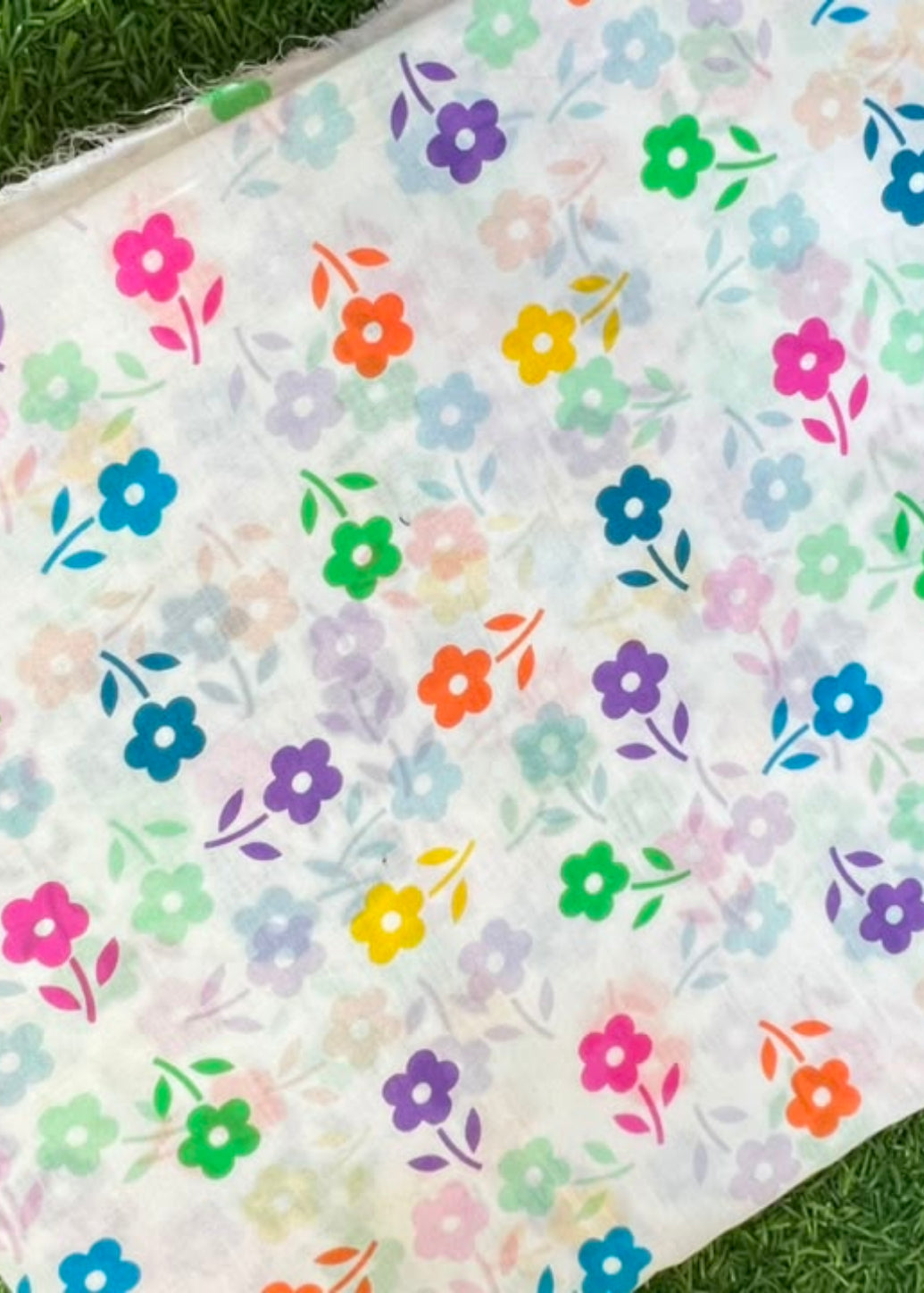 A rectangular piece of white fabric with a colorful all-over floral print, laid flat on green grass.