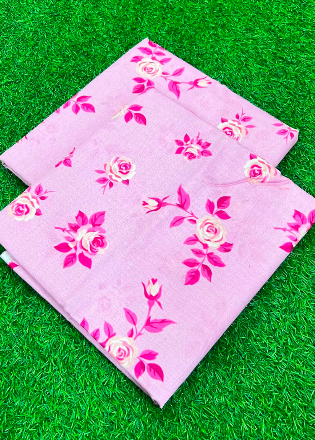 A folded light pink fabric with a pink and cream rose floral pattern, displayed on a green artificial grass background.
