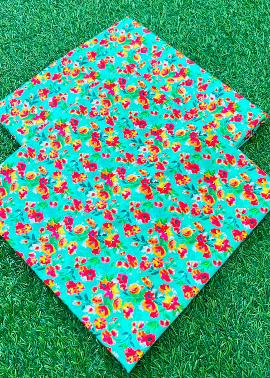 Two square mats with a vibrant floral pattern in red, yellow, and orange on a turquoise background, displayed on green grass.