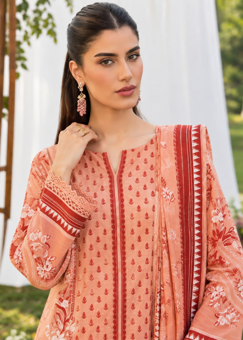 A woman wearing a peach-colored traditional embroidered kurta with red border details and a matching dupatta, photographed outdoors against a soft white draped backdrop.