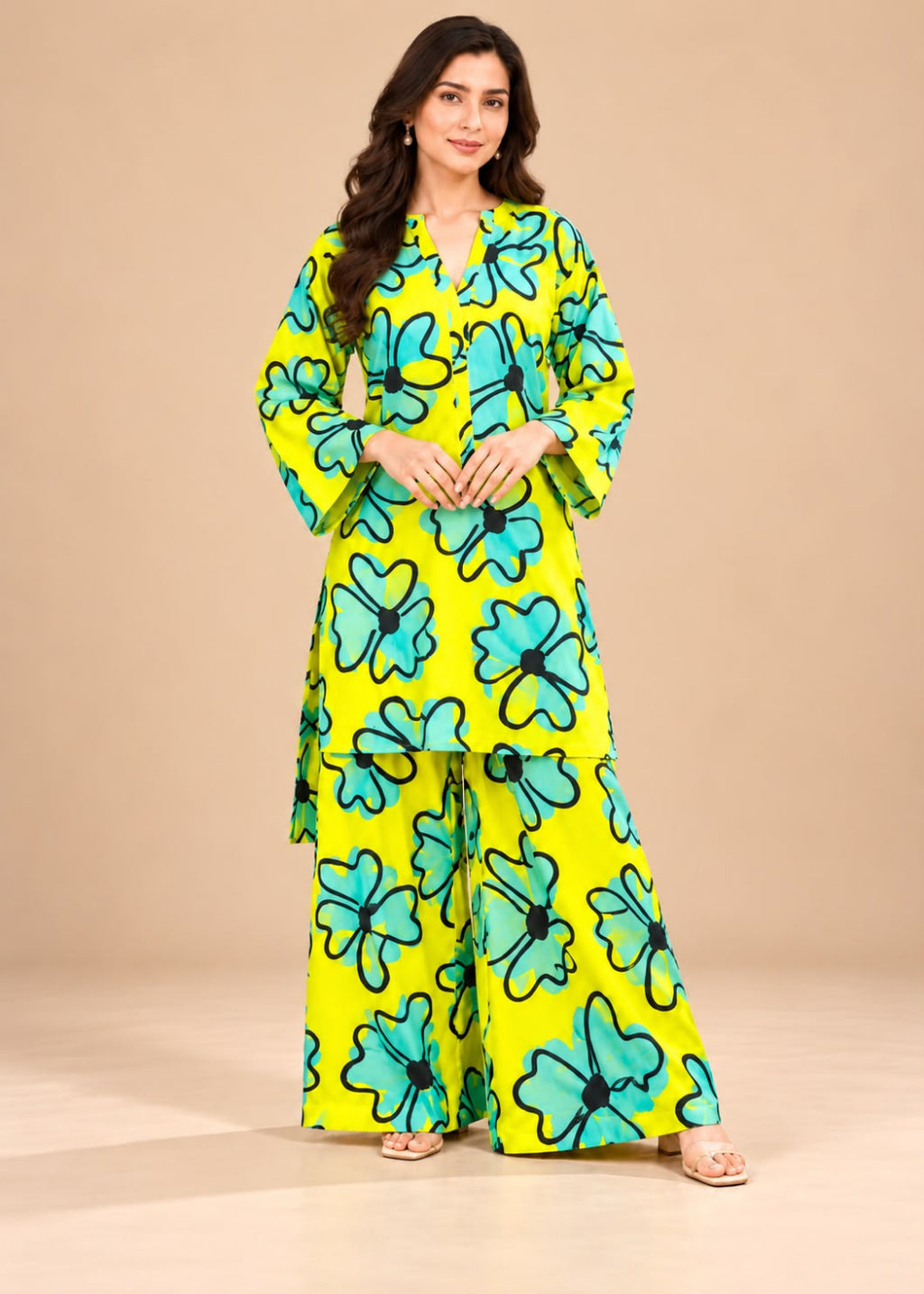 A woman modeling a bright yellow two-piece kurta set with a bold turquoise and black floral print, standing against a solid beige studio background.