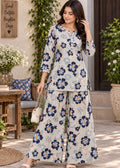 A woman models a two-piece floral print kurta and wide-leg trouser set in a styled outdoor patio setting. The set features a light beige base with navy blue and white floral motifs, accented with small yellow centers.