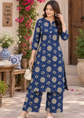 A woman models a navy blue three-piece kurta set with beige and white floral patterns, standing in a beautifully decorated outdoor patio setting with potted plants and wooden furniture.