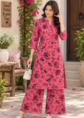 A woman models a three-piece pink floral kurta set with matching wide-leg pants and a small white handbag, standing in a garden courtyard setting.
