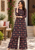 A woman models a dark navy blue three-piece kurta set with a colorful geometric and floral print, featuring a long kurta, wide-leg pants, and a matching belt, set against an outdoor patio backdrop.