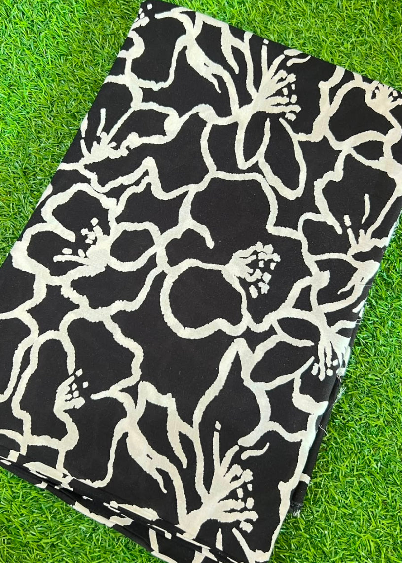A folded black fabric item, likely a blanket or towel, featuring a bold off-white floral pattern, laid flat on a bright green grass background.