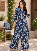 A woman models a navy blue three-piece kurta set with a light blue and white floral print, standing in a sunlit courtyard with potted plants and a wooden bench.