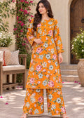 A woman models a bright orange floral print kurta and wide-leg pants set in an outdoor courtyard setting with stone flooring, potted plants, and a wooden door in the background.