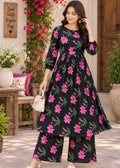 A woman models a black three-piece kurta set with a vibrant pink and green floral print, standing in a beautifully decorated outdoor patio setting.