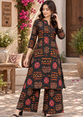 A woman models a black three-piece kurta set with a vibrant red, orange, and yellow geometric and floral pattern, set against a rustic outdoor patio backdrop.