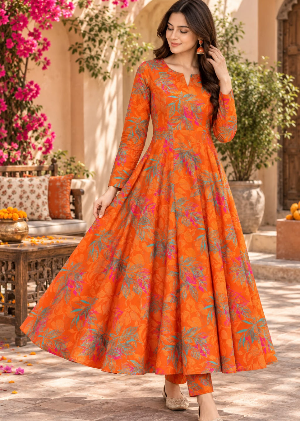 A woman models a vibrant orange floral print Anarkali gown with matching pants in an outdoor courtyard setting.