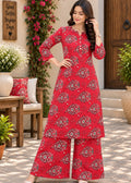 A woman models a red three-piece kurta set with a floral print, featuring a long-sleeve tunic, wide-leg pants, and a matching belt, set against an outdoor patio backdrop with plants and furniture.
