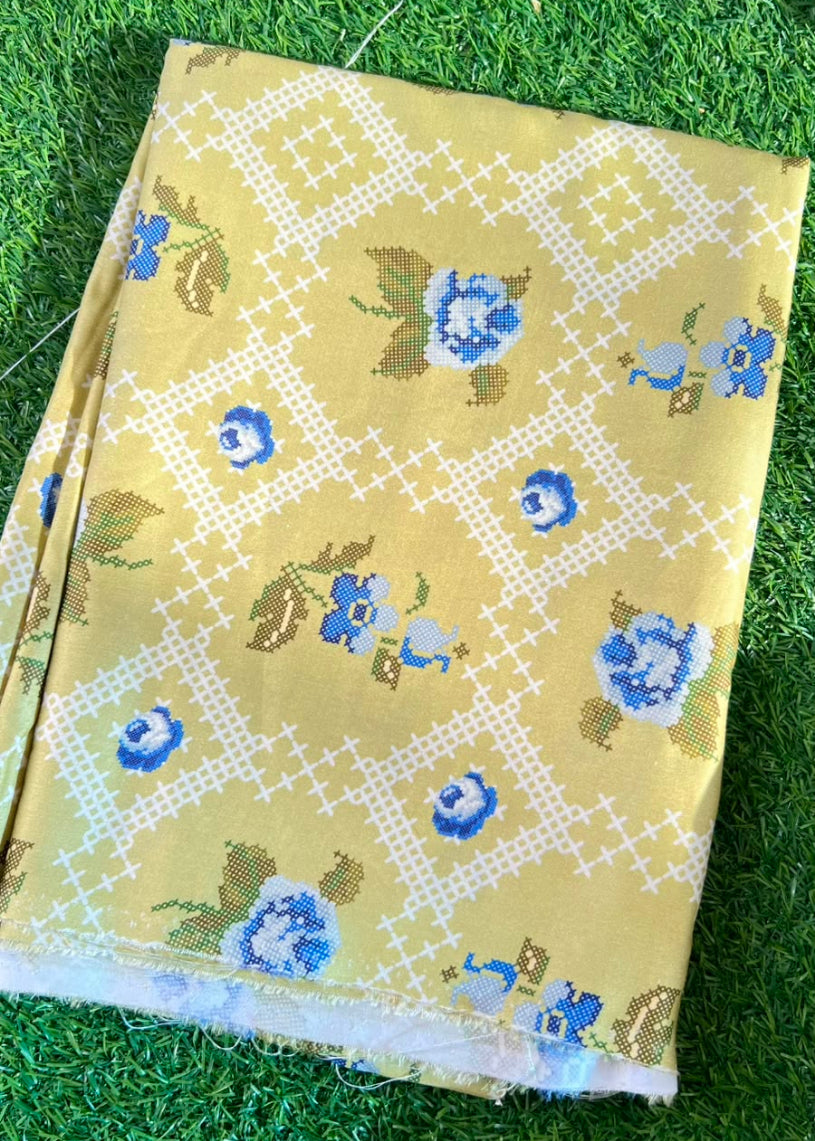 A folded piece of mustard yellow fabric with a white geometric lattice pattern and blue floral motifs, laid flat on green artificial grass.