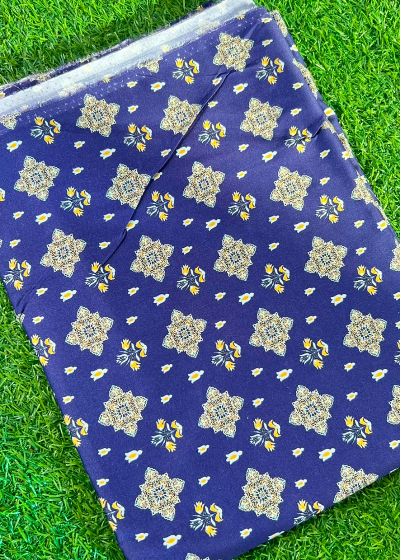 A folded piece of royal blue fabric with a beige and yellow floral mandala pattern, laid flat on green grass.