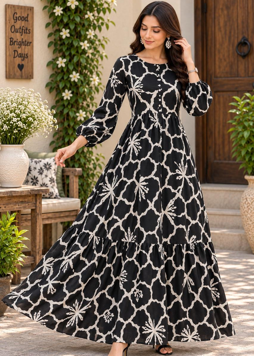 A woman modeling a long black maxi dress with a white geometric trellis pattern and floral motifs, standing in an outdoor patio setting.