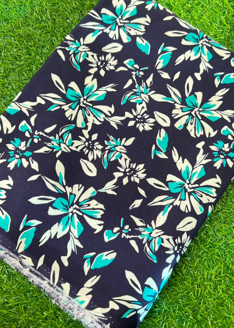 A rectangular textile or fabric sample with a black background and a repeating floral print in teal and cream, laid diagonally on bright green grass.