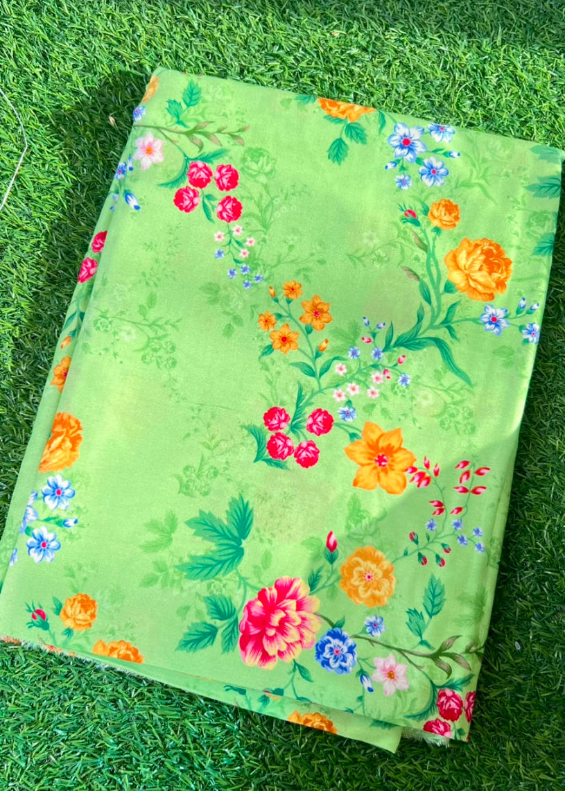 A folded light green fabric, likely a saree or textile, featuring a vibrant multicolor floral print with pink, orange, and blue flowers, laid flat on green grass.