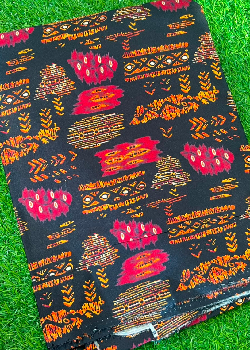 A folded piece of black fabric with a vibrant red, orange, and white geometric tribal pattern, displayed on a green grass background.
