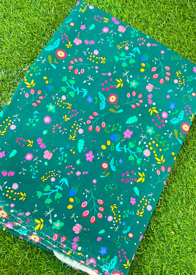 A rectangular piece of teal fabric with a colorful floral and botanical print, laid flat on green grass.
