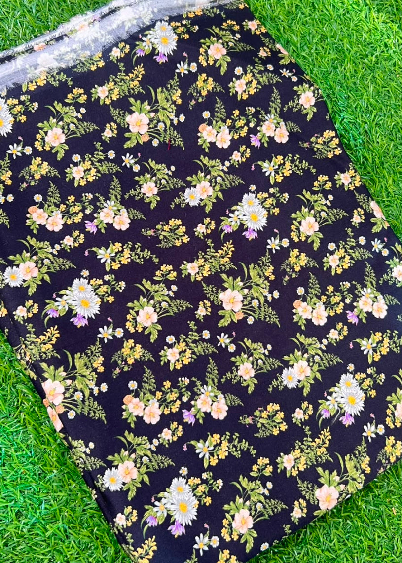 A folded black fabric item, likely a blanket or throw, featuring a dense floral print with white, yellow, and pink flowers and green leaves, laid flat on a green grass background.
