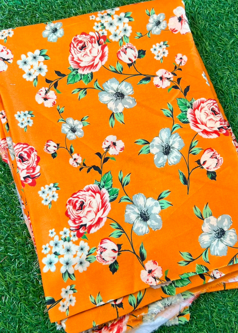A folded piece of bright orange fabric with a floral print, laid on green grass. The pattern features large pink and white roses, light blue flowers, and green leaves.