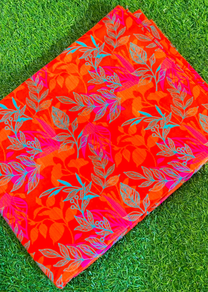 A folded red and orange fabric, likely a saree or scarf, featuring a vibrant floral and leaf pattern in pink, orange, and teal, laid flat on green grass.