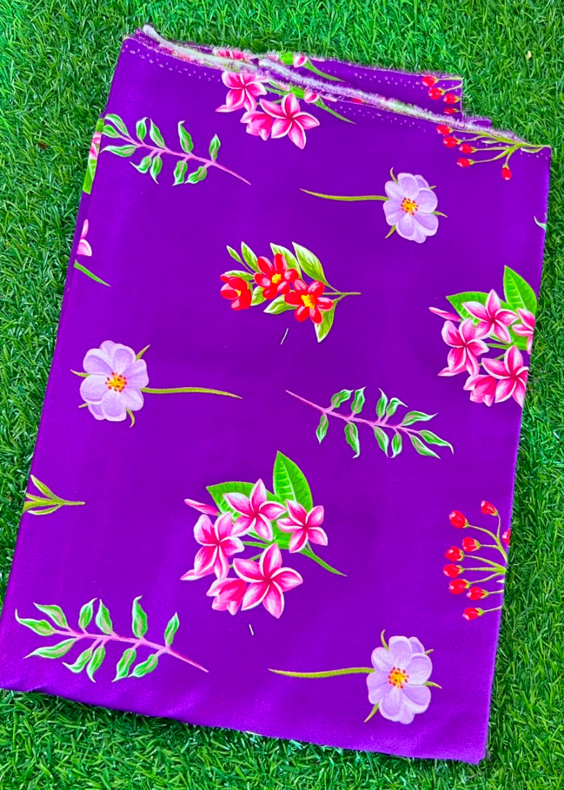 A folded purple fabric item, likely a pouch or small bag, featuring a colorful floral print with pink flowers, red berries, and green leaves, laid flat on a green grass background.