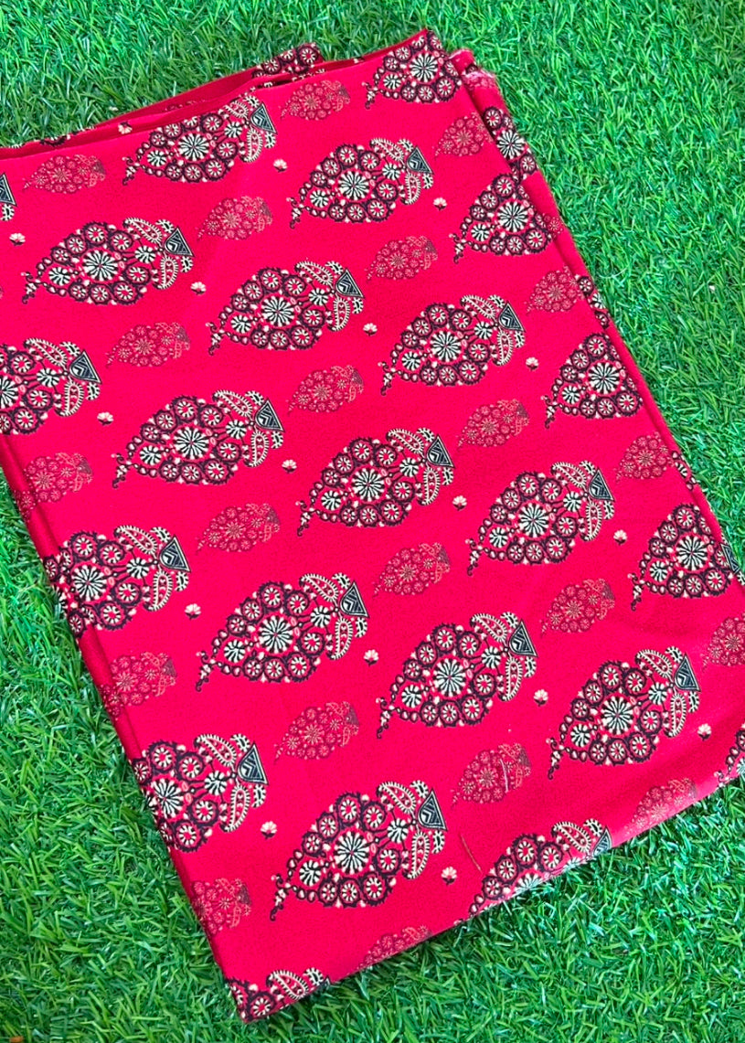 A folded bright pink fabric, likely a saree or scarf, featuring a detailed red, black, and white floral and paisley block print, laid flat on green grass.