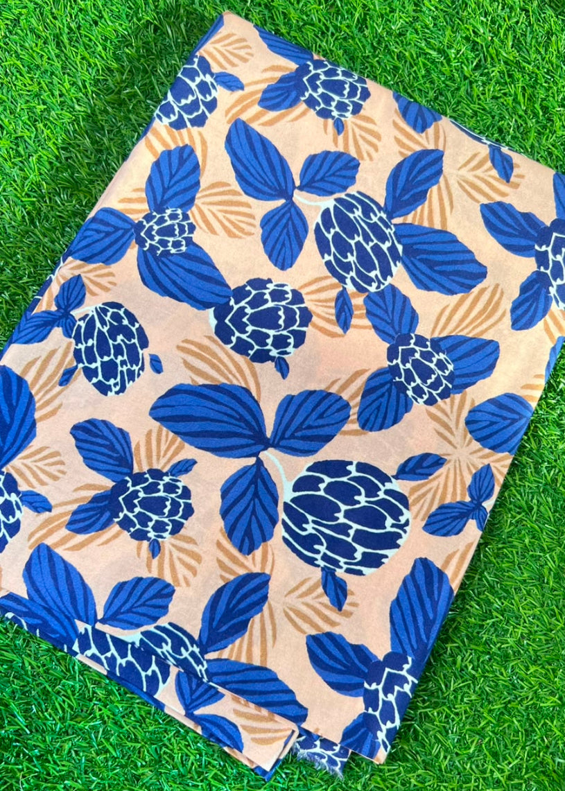 A folded piece of fabric with a blue and beige floral print, laid flat on green grass. A person's toe is visible in the bottom right corner.