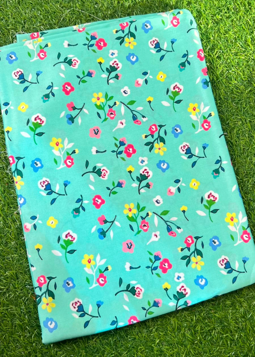 A folded mint green fabric item, likely a blanket or towel, featuring a colorful all-over floral print, laid flat on a green grass background.