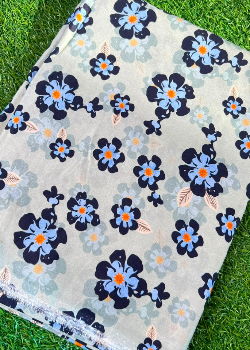 A folded piece of fabric with a floral print, laid flat on green grass. The fabric has a light beige background with dark navy blue flowers, light blue flowers, and small orange flower centers.