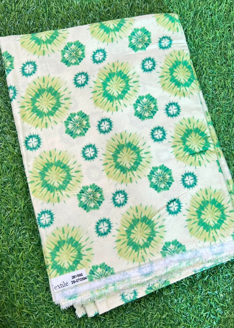 A folded cotton fabric with a green and yellow sunburst ikat pattern on a cream background, displayed on green grass. A white label is visible in the bottom left corner.