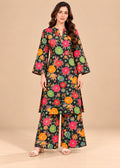 A woman modeling a black three-piece floral print kurta set with wide-leg pants, featuring colorful flowers in pink, orange, yellow, and green on a beige studio background.
