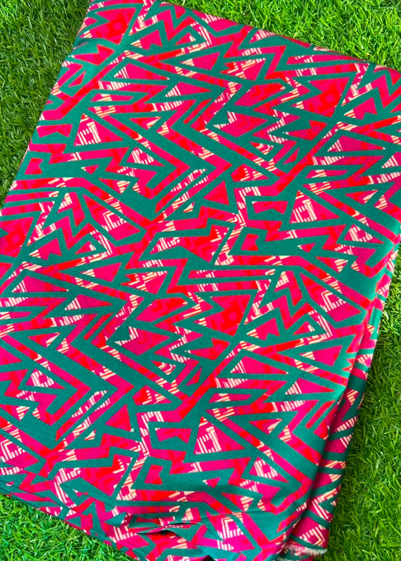 A folded piece of vibrant pink and green patterned fabric, likely a cotton sarje or textile, featuring a bold geometric zigzag design, laid flat on a green grass background.