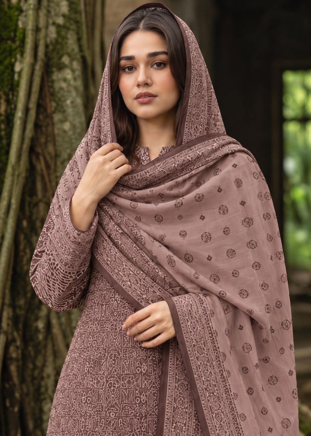 A woman wearing a dusty mauve printed salwar suit with a matching dupatta, standing outdoors in front of a tree trunk.