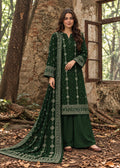 A woman stands outdoors in front of a rustic stone wall and window, wearing a dark green traditional outfit with white embroidered details and a matching dupatta.
