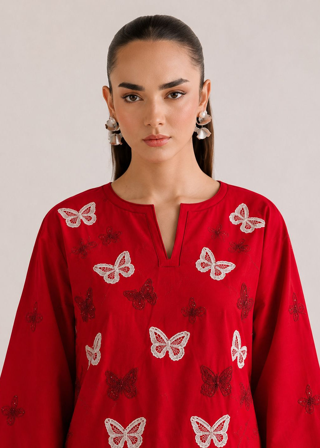 A woman wearing a bright red long-sleeve tunic top with embroidered white and dark red butterfly motifs, photographed against a plain light gray studio background.