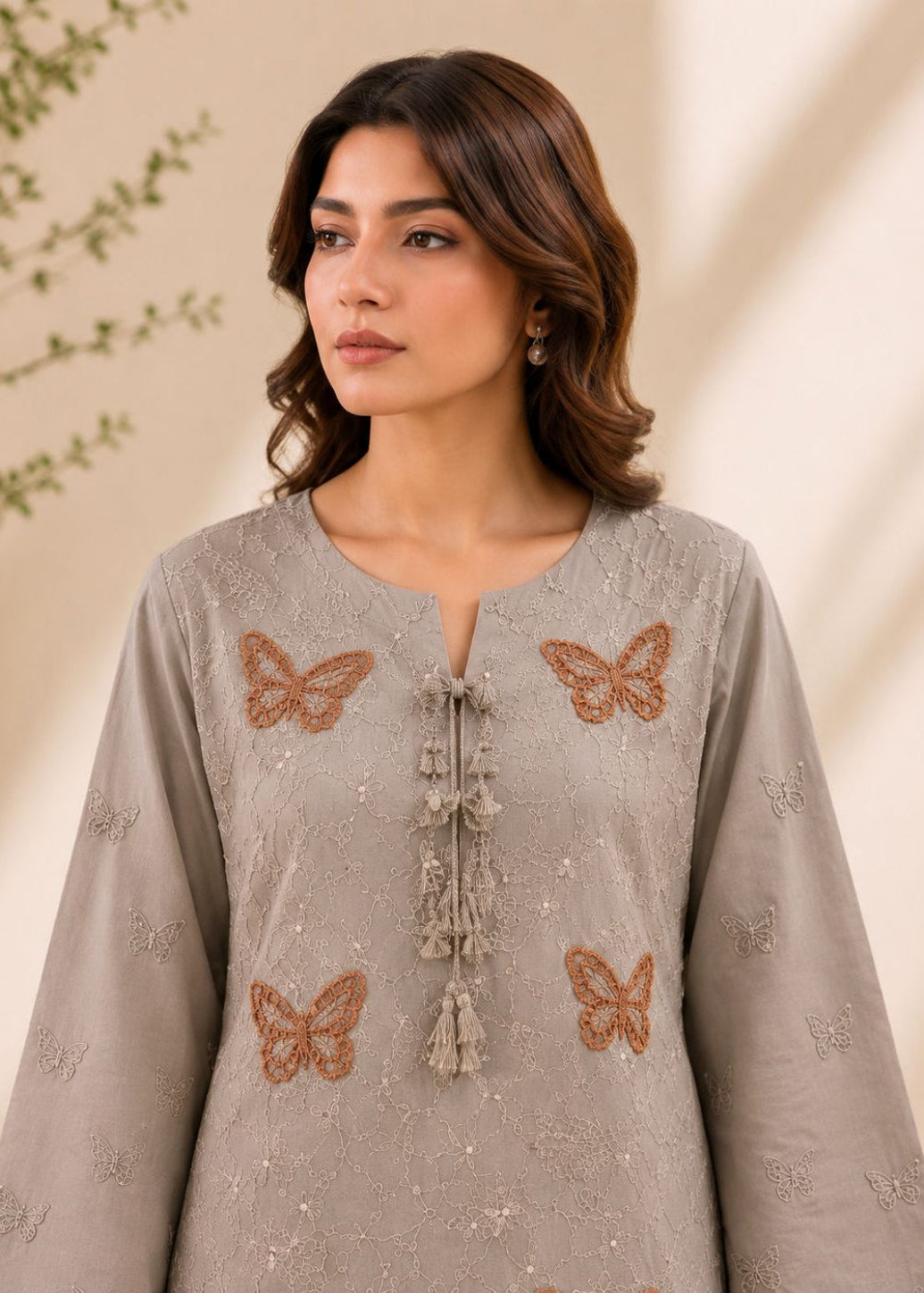 A woman wearing a light taupe-gray long-sleeve tunic with brown butterfly embroidery and tassel tie details, set against a warm beige background with soft leaf shadows.