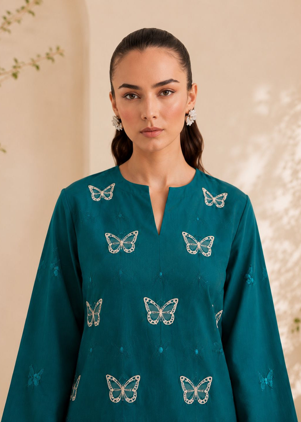A woman wearing a teal long-sleeve kurta with embroidered butterfly motifs, photographed against a warm beige background with soft leaf shadows.