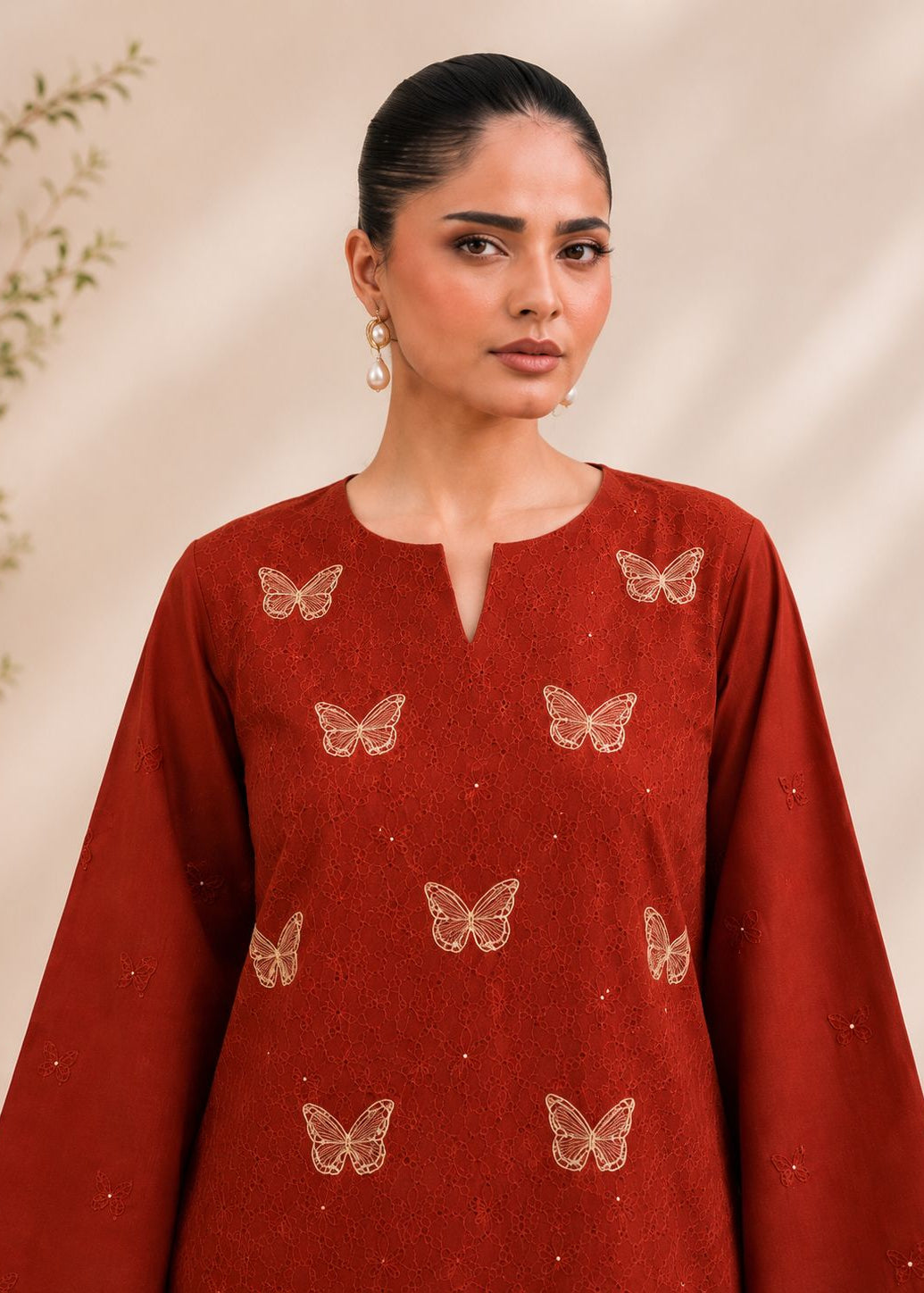 A woman wearing a rust-red long-sleeve kurta with gold butterfly embroidery, set against a warm beige background with soft leaf shadows.