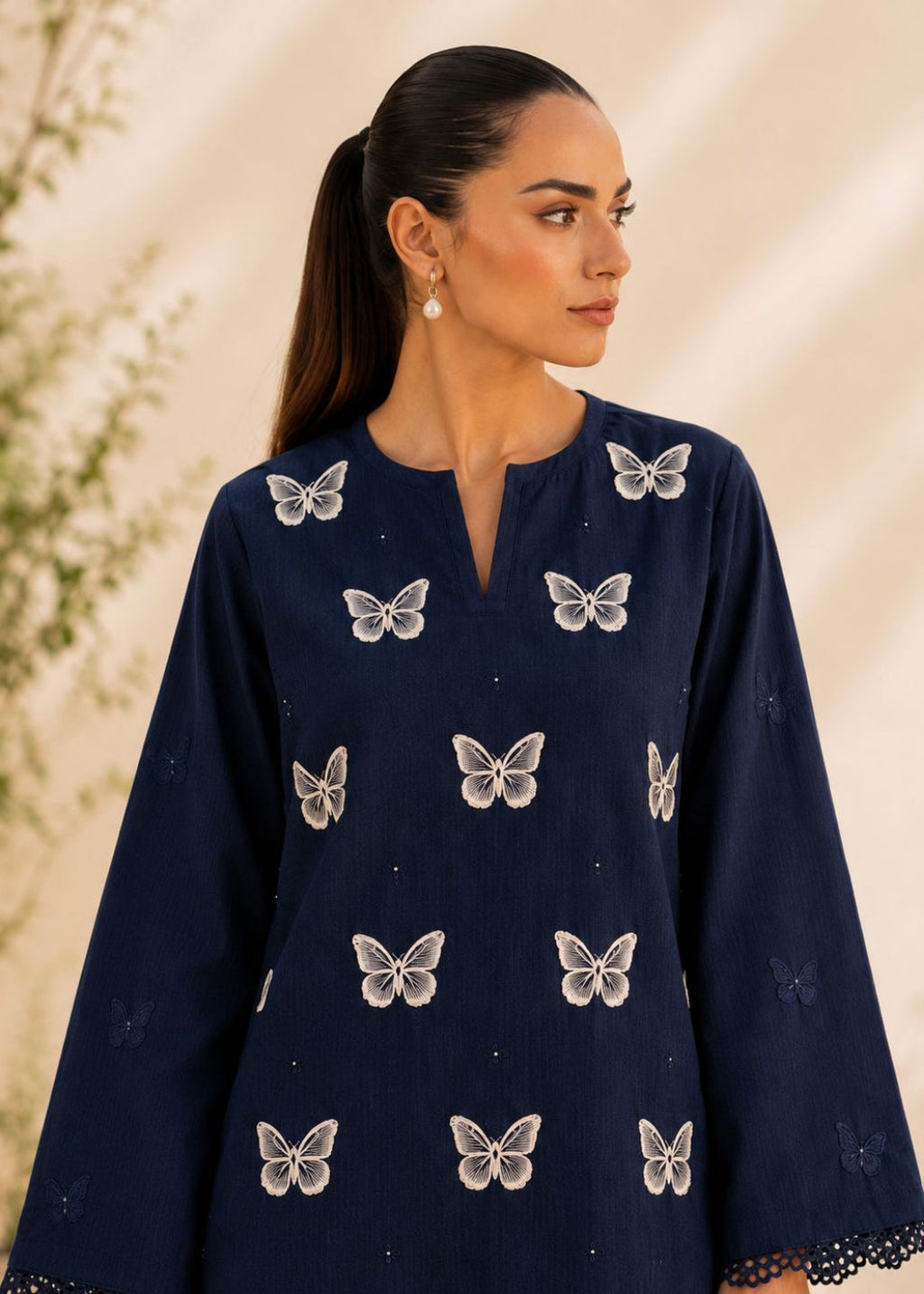 A woman wearing a navy blue long-sleeve kurta with white embroidered butterfly motifs, shown in a lifestyle setting with a warm beige background.