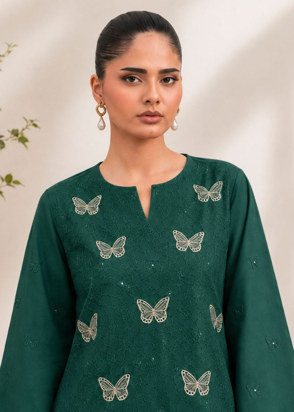 A woman wearing a dark green kurta with embroidered butterfly motifs, photographed against a soft beige background with subtle leafy decor at the side.