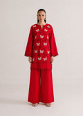 A model stands against a plain light beige studio background wearing a bright red traditional outfit consisting of a long-sleeve embroidered kurta with butterfly motifs and matching wide-leg trousers.